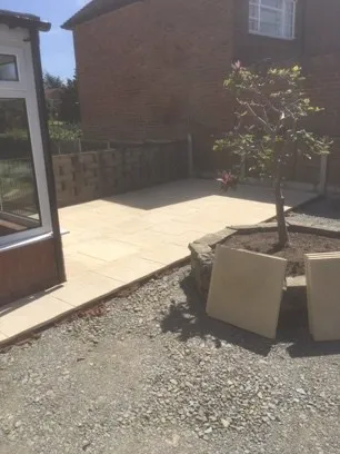 A partially completed light coloured slabbed patio contrasting with grey hardcore. A tree stands close by