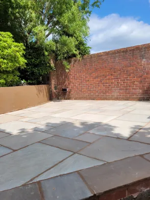 A light coloured slabbed patio contrasts with a red wall, blue sky and bright green vegetation